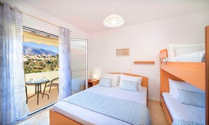 A bedroom with a bed, a bedside table with a lamp, a view of a village through a window, and a balcony with a table and chairs. - Villa di Palma Heated Pool (Georgioupolis)
