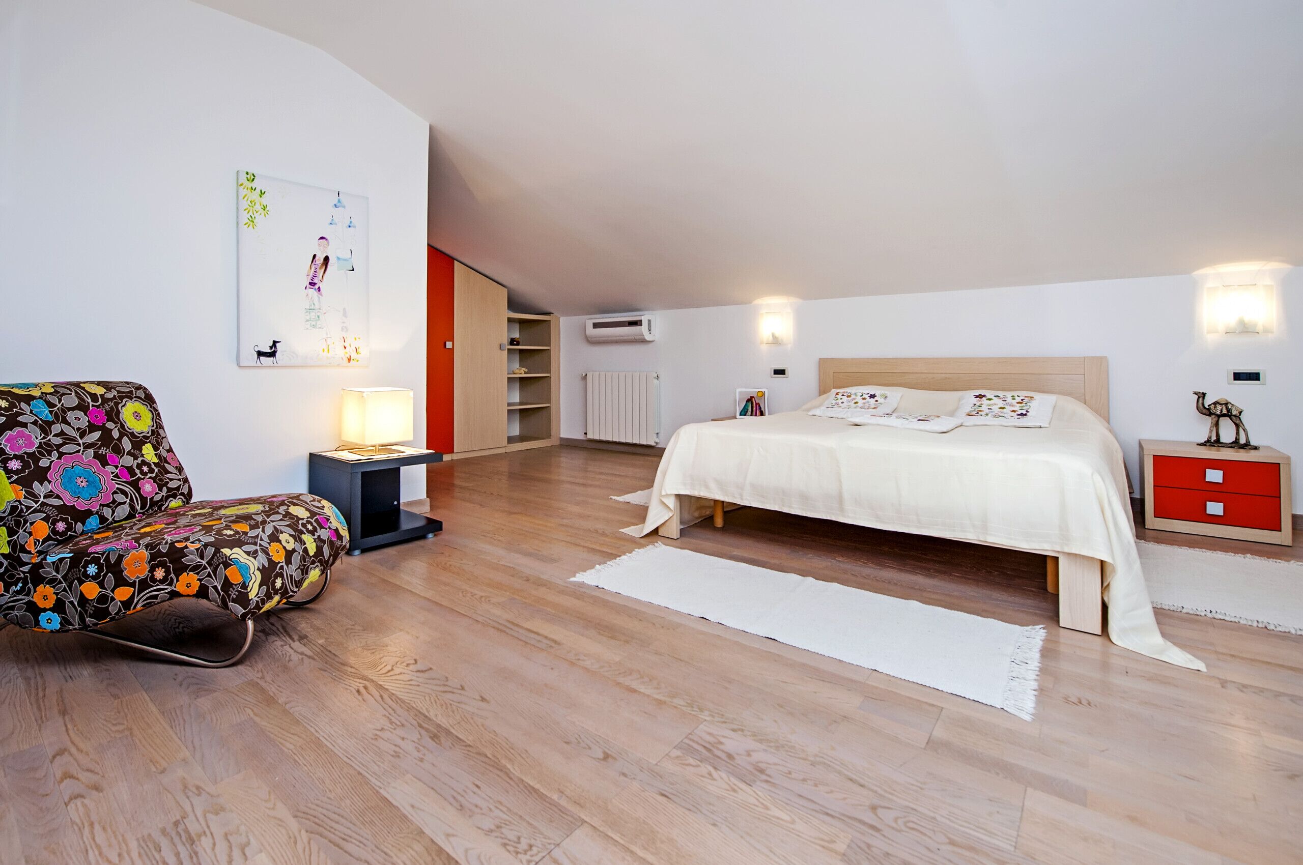 A bedroom with a bed, a chair, a nightstand, a lamp, a radiator, and a wall-mounted shelf.