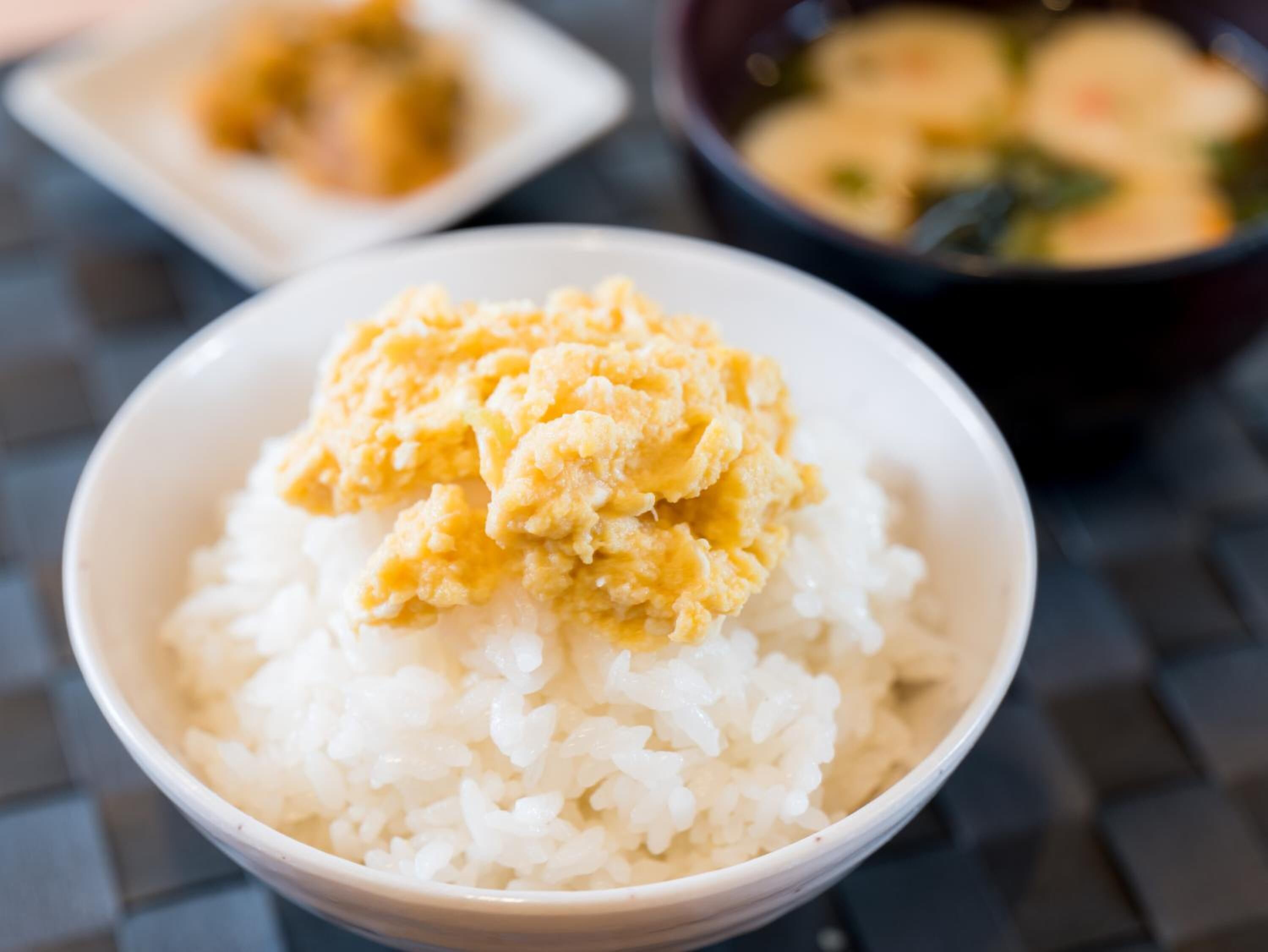 A bowl of white rice topped with scrambled eggs, with a bowl of soup in the background.
