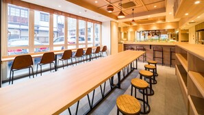 A modern dining area with a long table and several stools, large windows with a view of the street, and a kitchen area with a bar counter. - Super Hotel Hirosaki (Hirosaki)