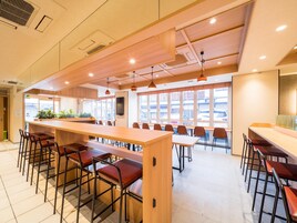 A spacious dining area with long wooden tables and metal chairs, large windows with a view of the street, and a modern ceiling design.