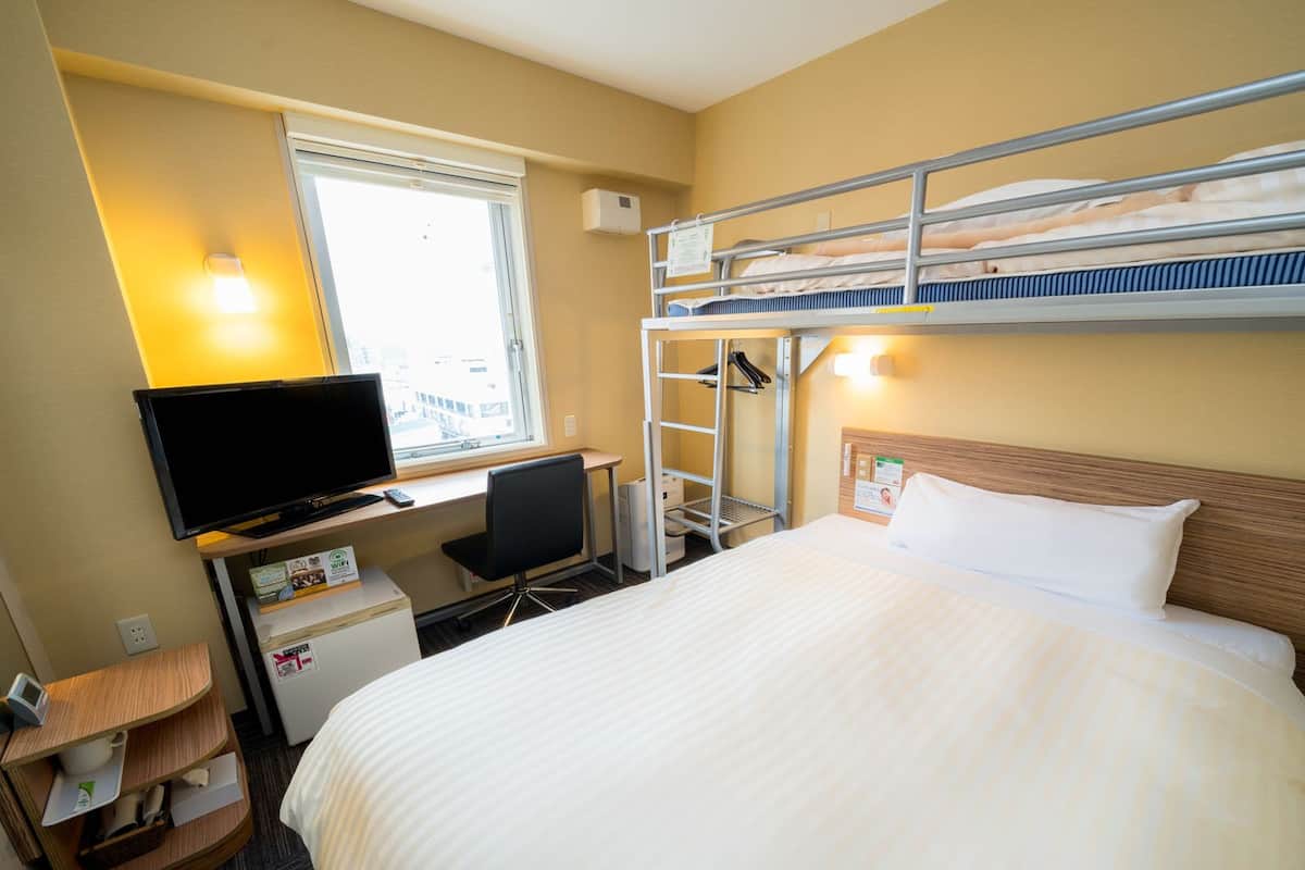 A hotel room with a bunk bed, a desk with a computer, a television, and a small refrigerator.