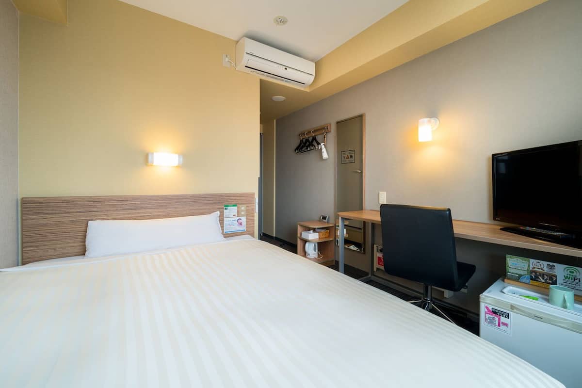 A hotel room with a bed, a desk, a chair, a television, and an air conditioning unit.