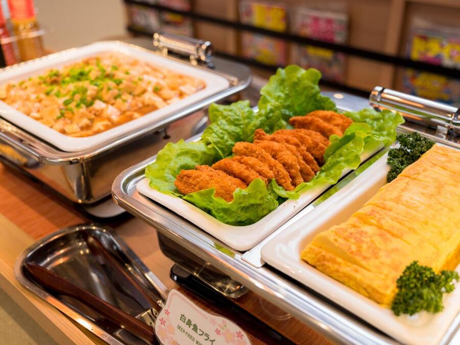 Ein Buffet mit verschiedenen Speisen, darunter frittierte Gerichte, eine Auflaufform und eine Schichtspeise, alles serviert auf Metalltabletts.