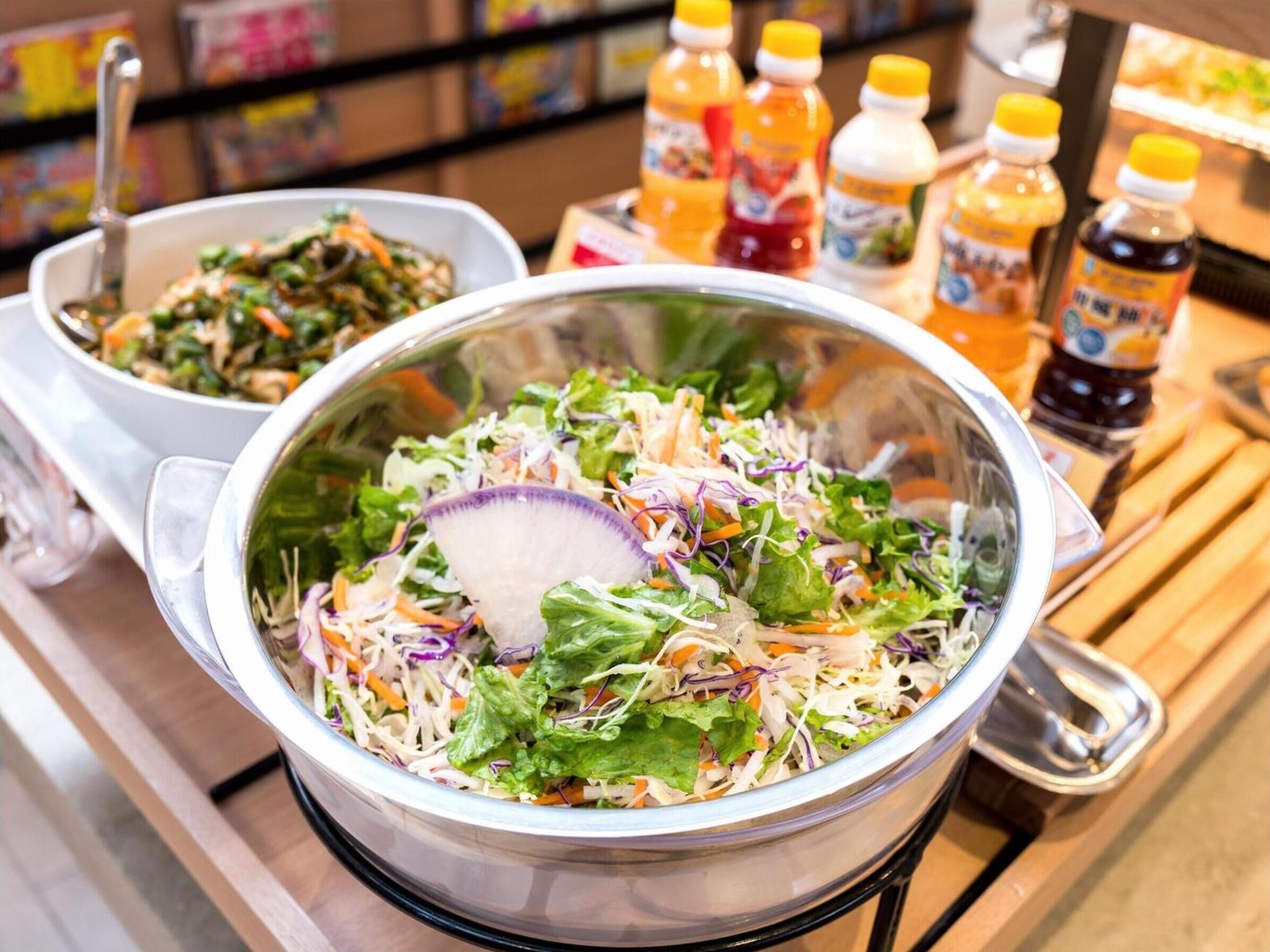 Un tazón de ensalada con diversos vegetales y una cuchara para aderezo, rodeado de botellas de condimentos.