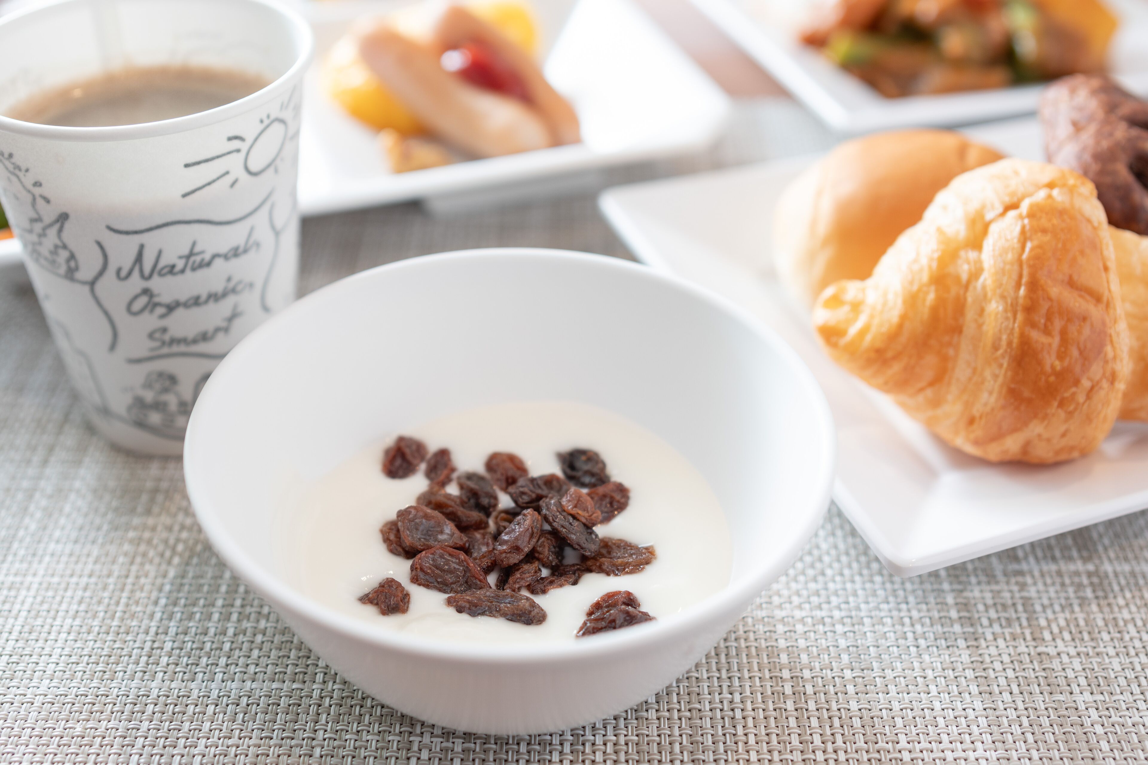 Une table mise pour le petit-déjeuner, avec un bol de yaourt et de raisins secs, un croissant et une tasse de café.