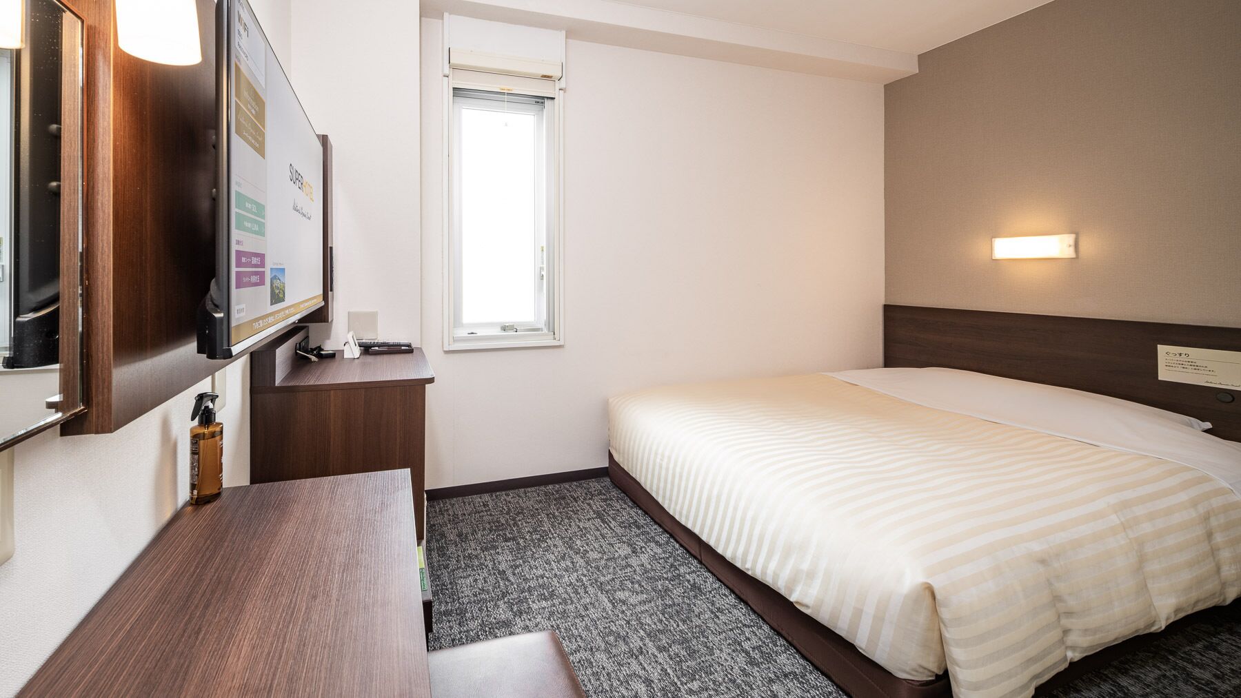 Ett hotellrum med en säng, ett skrivbord, en tv, ett fönster och en vägglampa.
