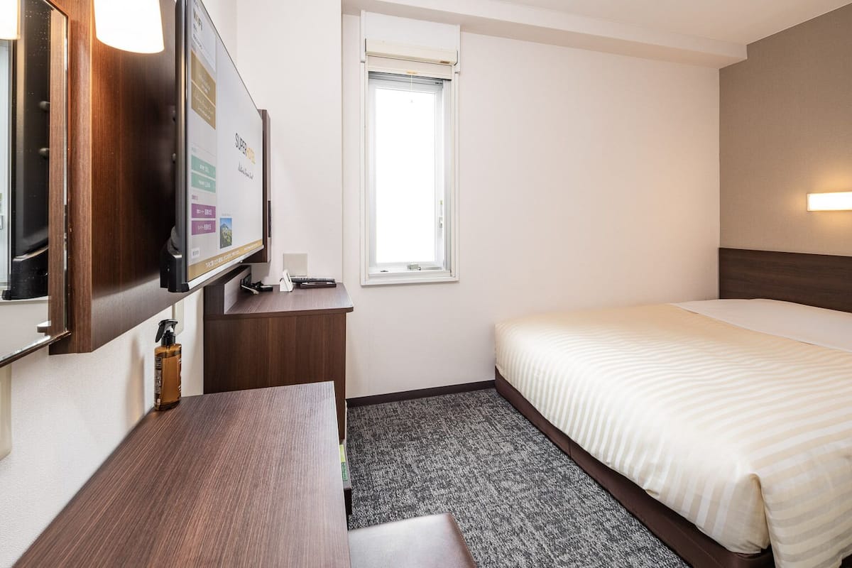 Habitació d'hotel amb un llit, una taula de nit, una televisió, una finestra i una làmpada penjada a la paret.