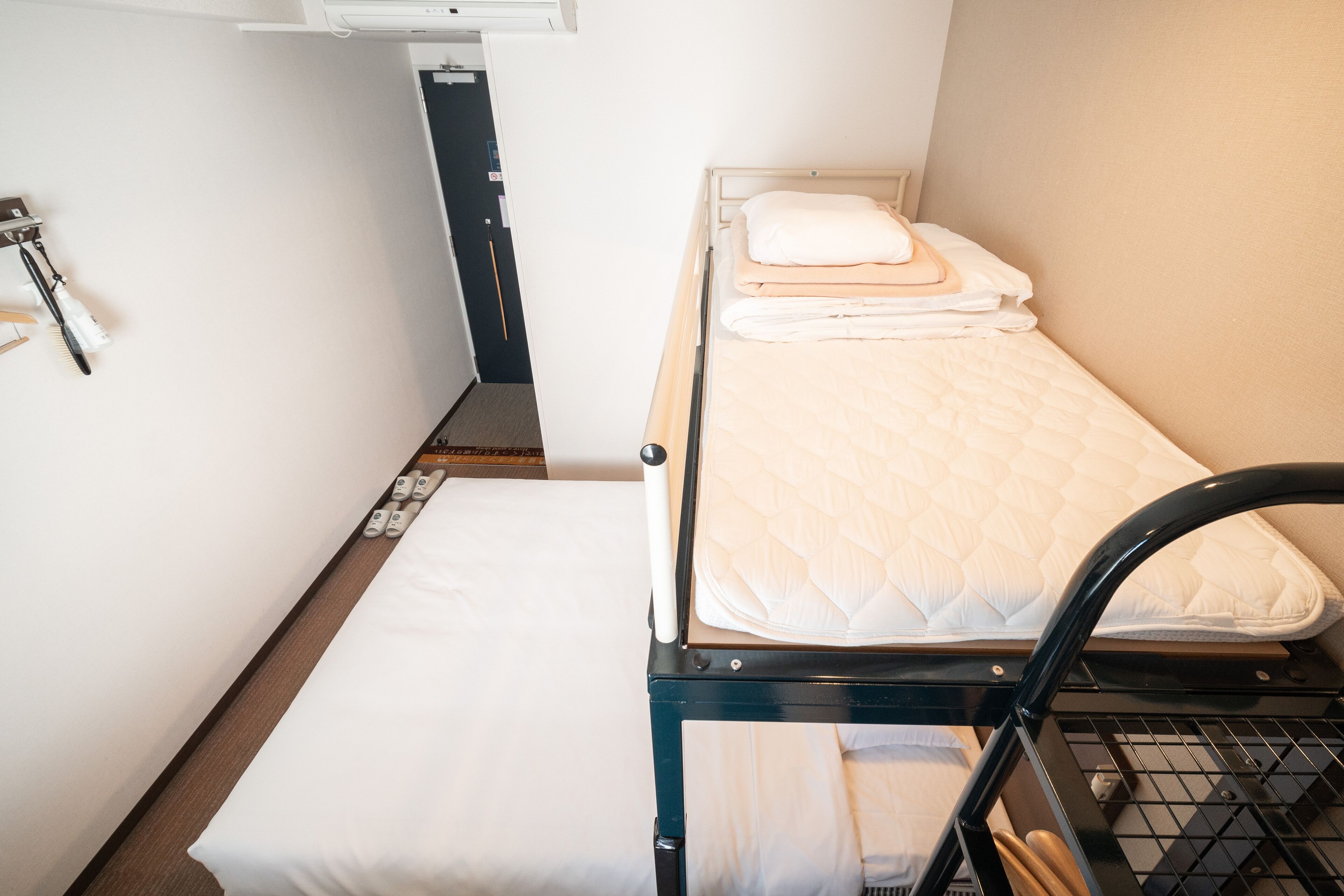 A small, single-bed room with a wooden frame and a mattress. There is a small table with a chair and a wall-mounted shelf with a bag.