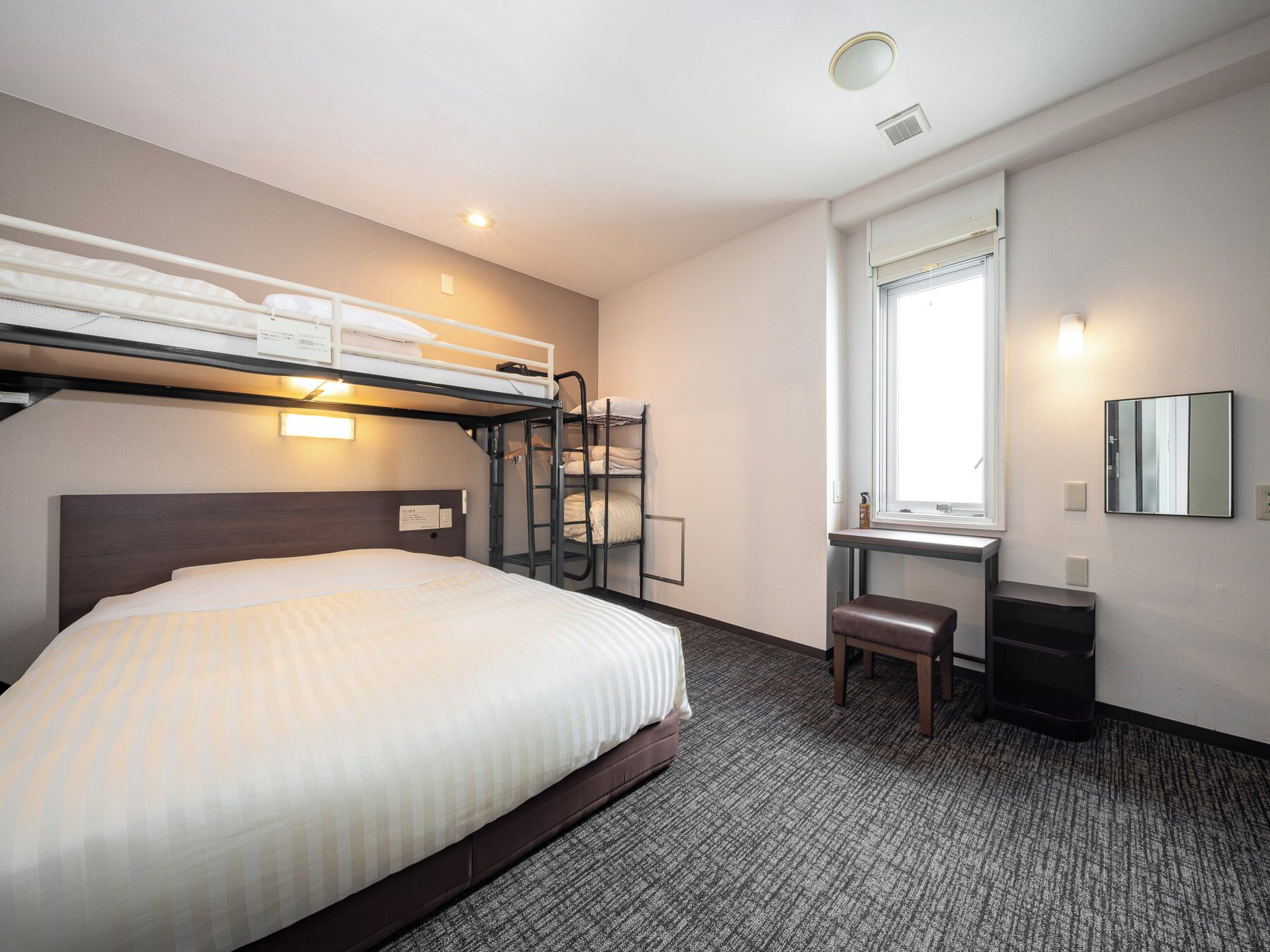 A hotel room with a bunk bed, a desk, a chair, a mirror, and a window.