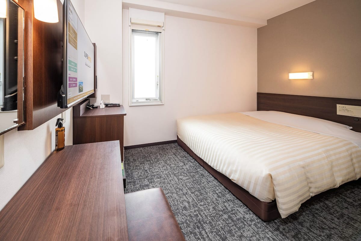 Habitació d'hotel amb un llit, escriptori, televisió, finestra i una làmpada penjada a la paret.