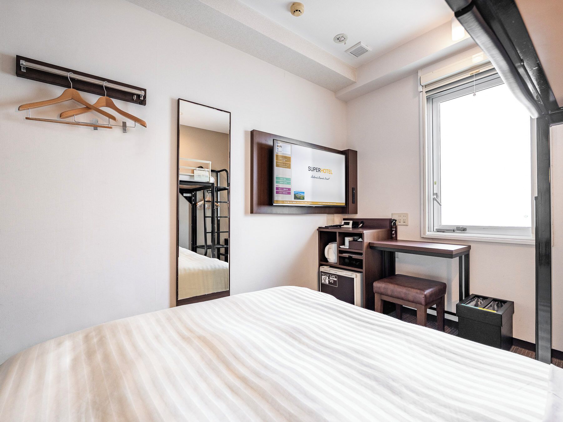 A hotel room with a bed, a desk with a computer, a mirror, a TV, and a window.