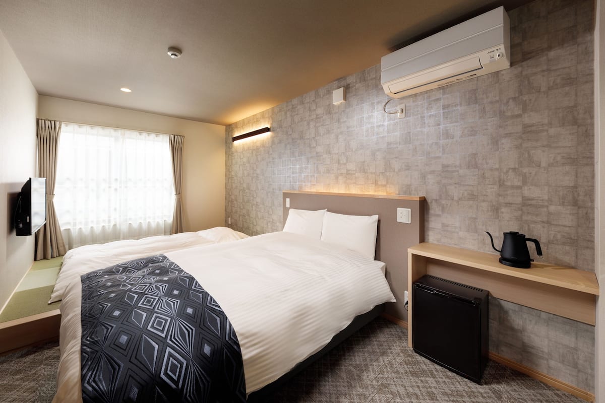 A hotel room with a bed, a small desk with a kettle, an air conditioner, and a view of the outdoors through a window with curtains.