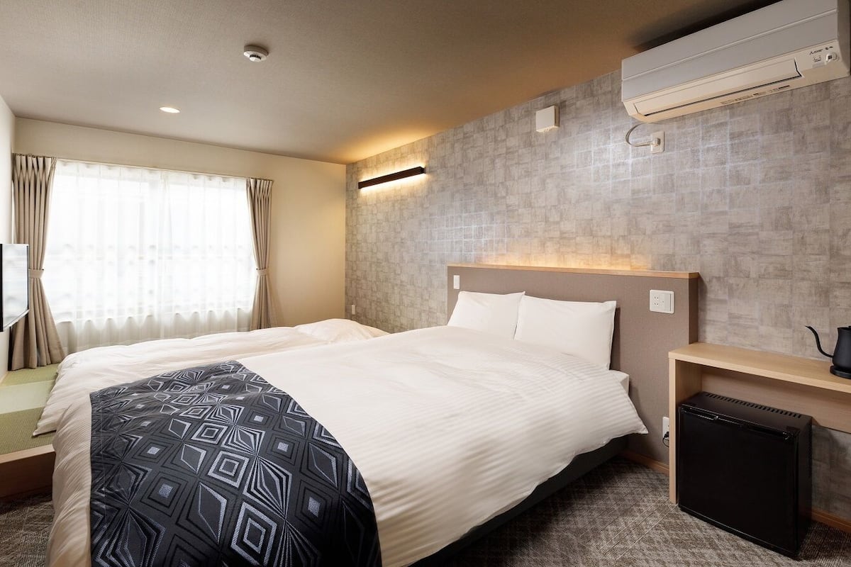 A hotel room with a bed, a bedside table with a kettle, a wall-mounted air conditioner, and a window with curtains.