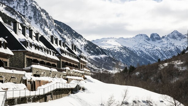 Filera d'edificis de pedra amb terrasses cobertes de neu, davant d'un paisatge de muntanyes nevades.