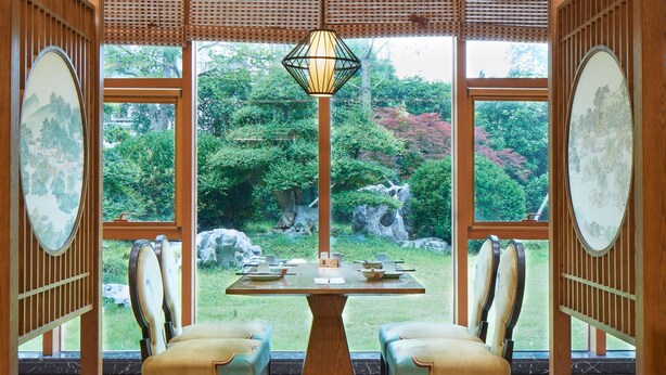 Habitación de estilo tradicional con una mesa de comedor, dos sillas y una lámpara colgante. Amplias ventanas ofrecen vistas a un jardín con rocas y árboles.