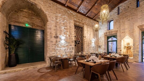 Een restaurant met rustieke stenen muren, houten tafels en stoelen, en een hoog plafond met zichtbare balken.
