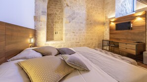 Quarto com parede de pedra, uma escrivaninha de madeira, televisão e cama com almofadas estampadas.