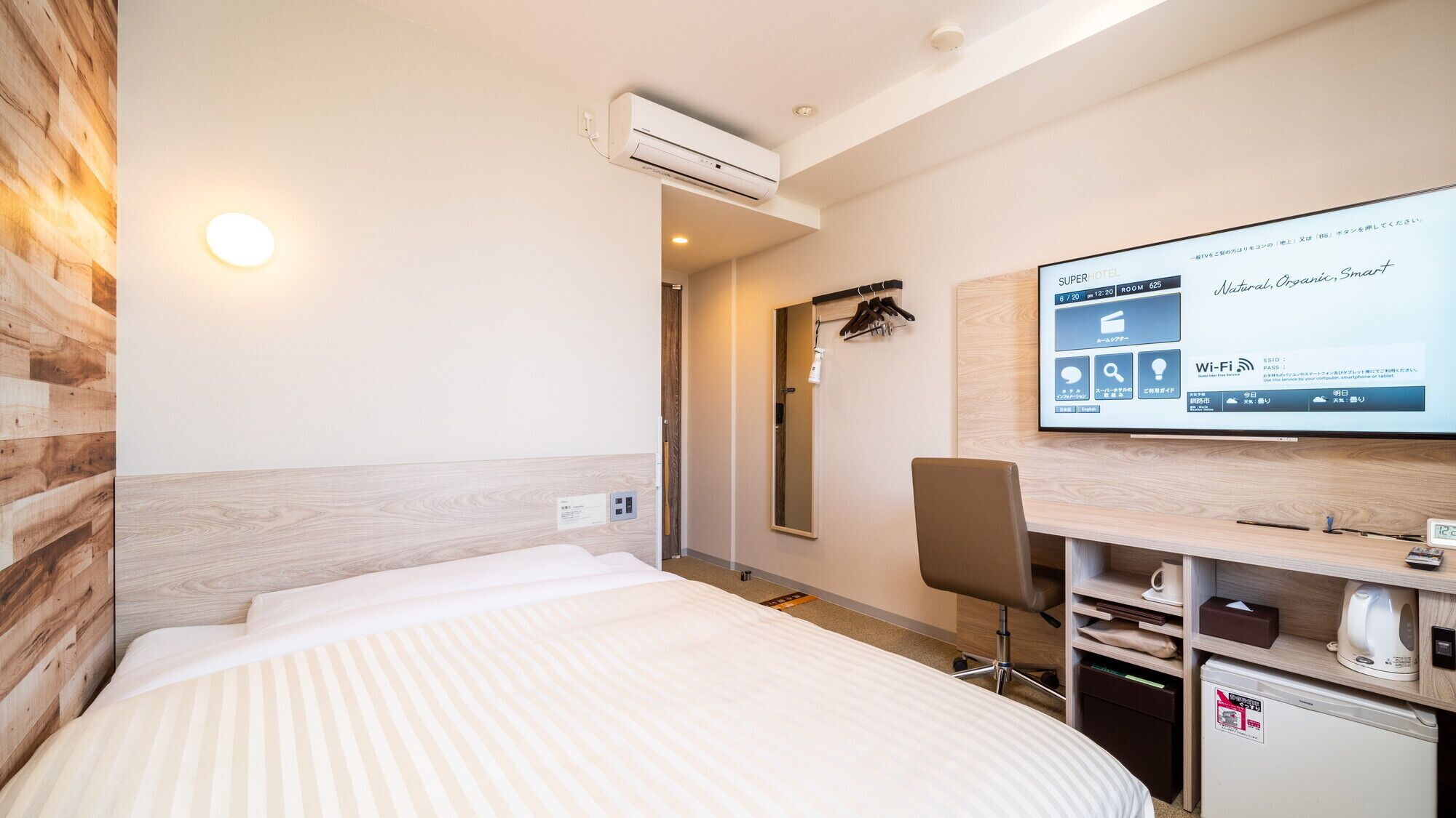 Une chambre d’hôtel équipée d’un lit, d’un bureau avec un ordinateur, d’un climatiseur et d’un téléviseur affichant des informations sur l’hôtel.