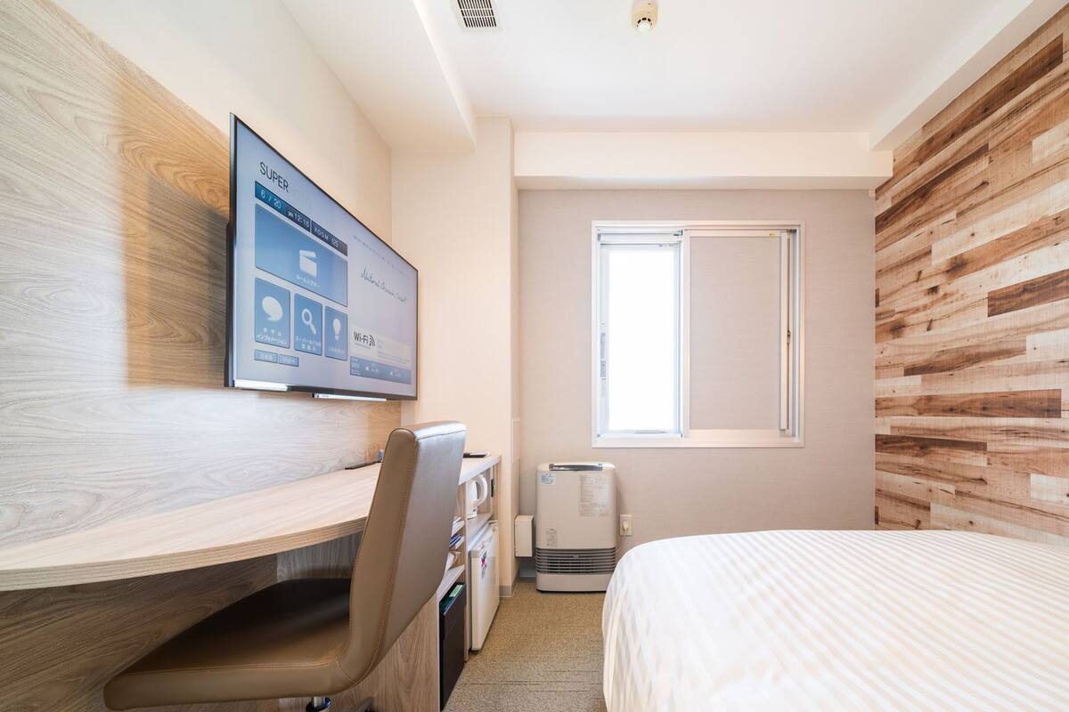 A hotel room with a wooden desk, a bed, a TV mounted on the wall, and a window.