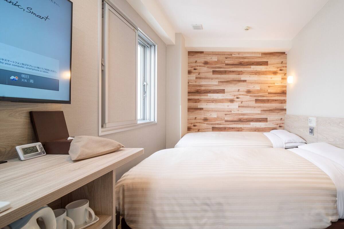 A modern hotel room with a wooden wall panel, a bed with white bedding, a desk with a laptop, and a digital clock.