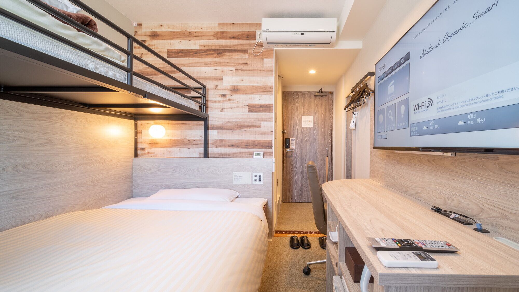 Une chambre d’hôtel compacte équipée d’un lit superposé, d’un bureau avec un ordinateur portable et d’un grand écran affichant des informations sur la chambre.