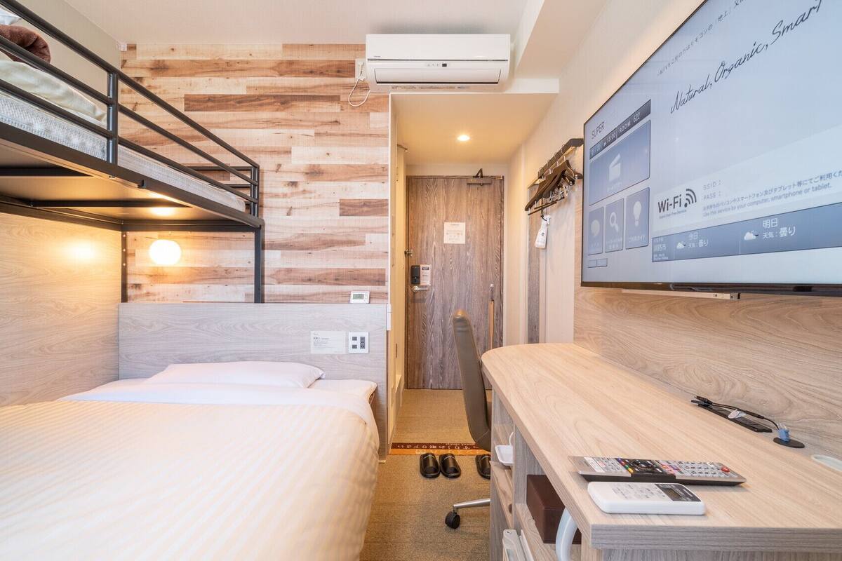 A compact hotel room with a bunk bed, a desk with a laptop, and a large screen displaying room information.