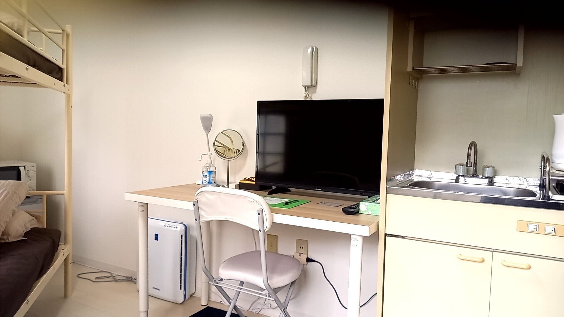 A compact room with a bunk bed, a desk with a computer, a chair, a small fridge, a microwave, a sink, and a mirror.