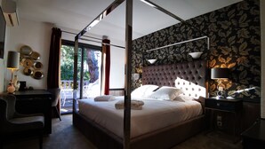 A four-poster bed with a tufted headboard, a desk with a lamp, and a chair in a room with floral wallpaper and a view of greenery outside. - La Villa Dune, Hôtel & Spa  (Gassin)
