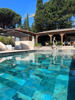 A clear blue pool with tiled flooring, outdoor seating under a pergola, and a view of trees and a distant construction crane. - La Villa Dune, Hôtel & Spa  (Gassin)