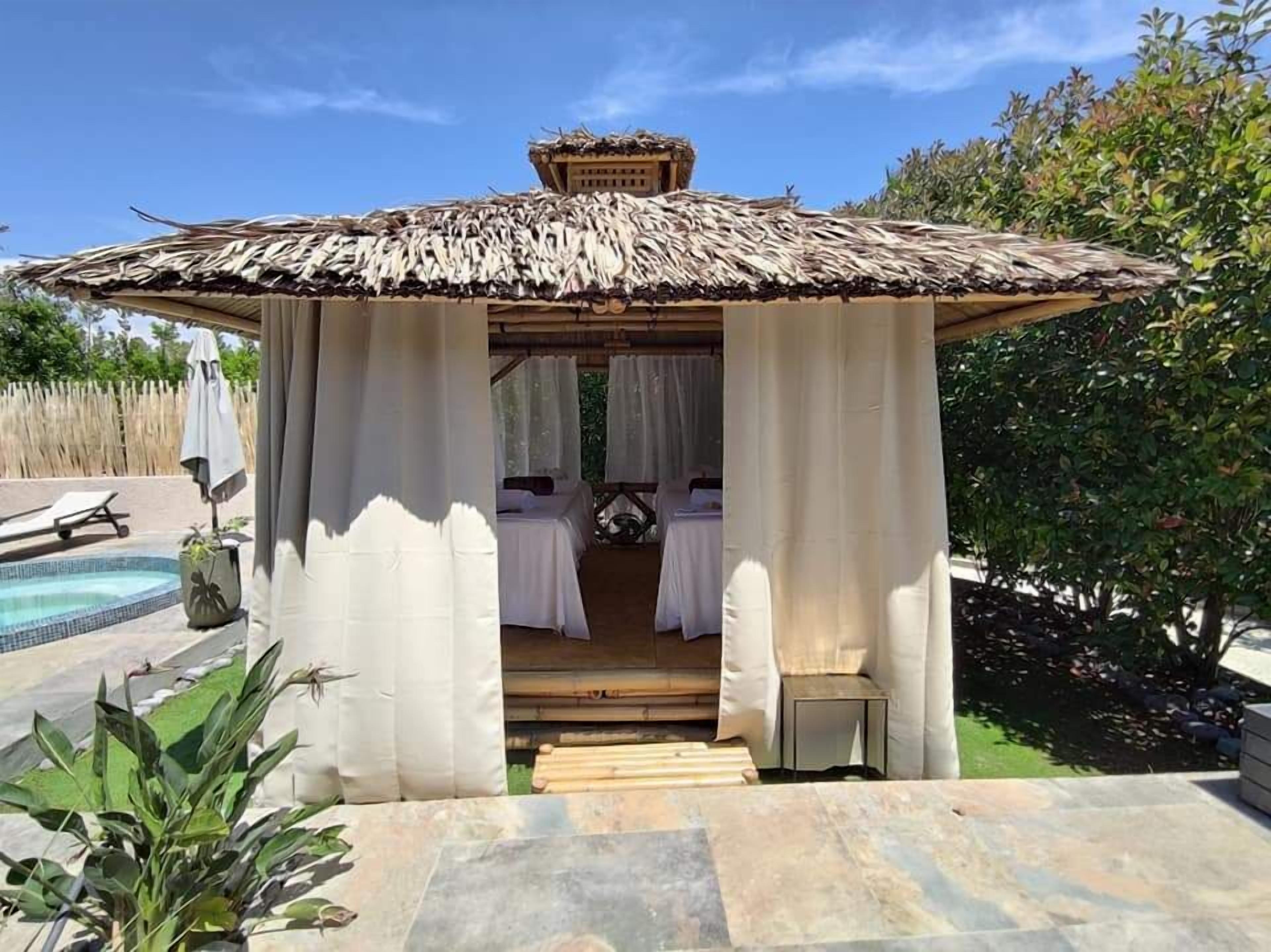 Un belvédère au toit de chaume, agrémenté de rideaux blancs, avec une piscine et un jardin.