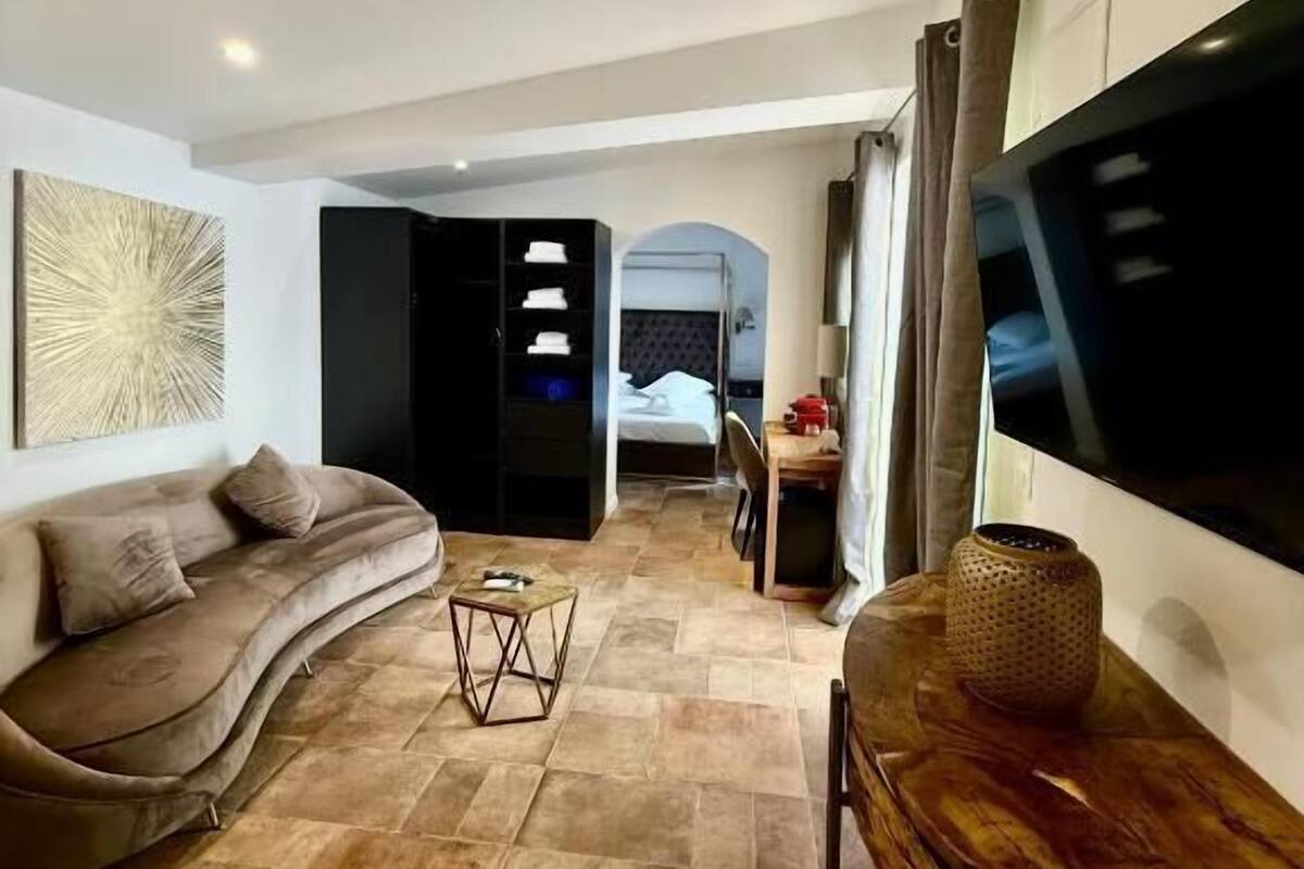 Un salon moderne avec un canapé, une table basse et un téléviseur. On aperçoit un lit au fond et une porte qui donne sur une autre pièce.