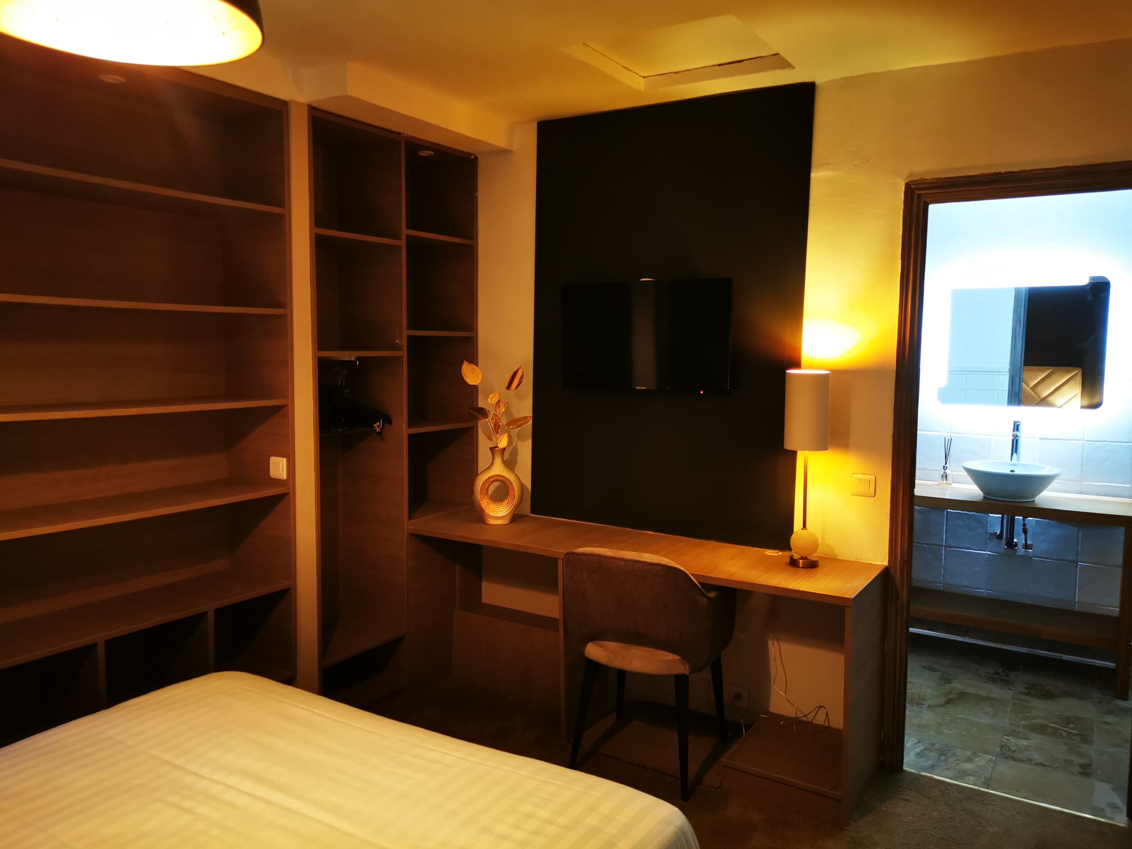 A hotel room with a bed, a desk with a chair, a TV, and a bathroom with a sink and mirror.