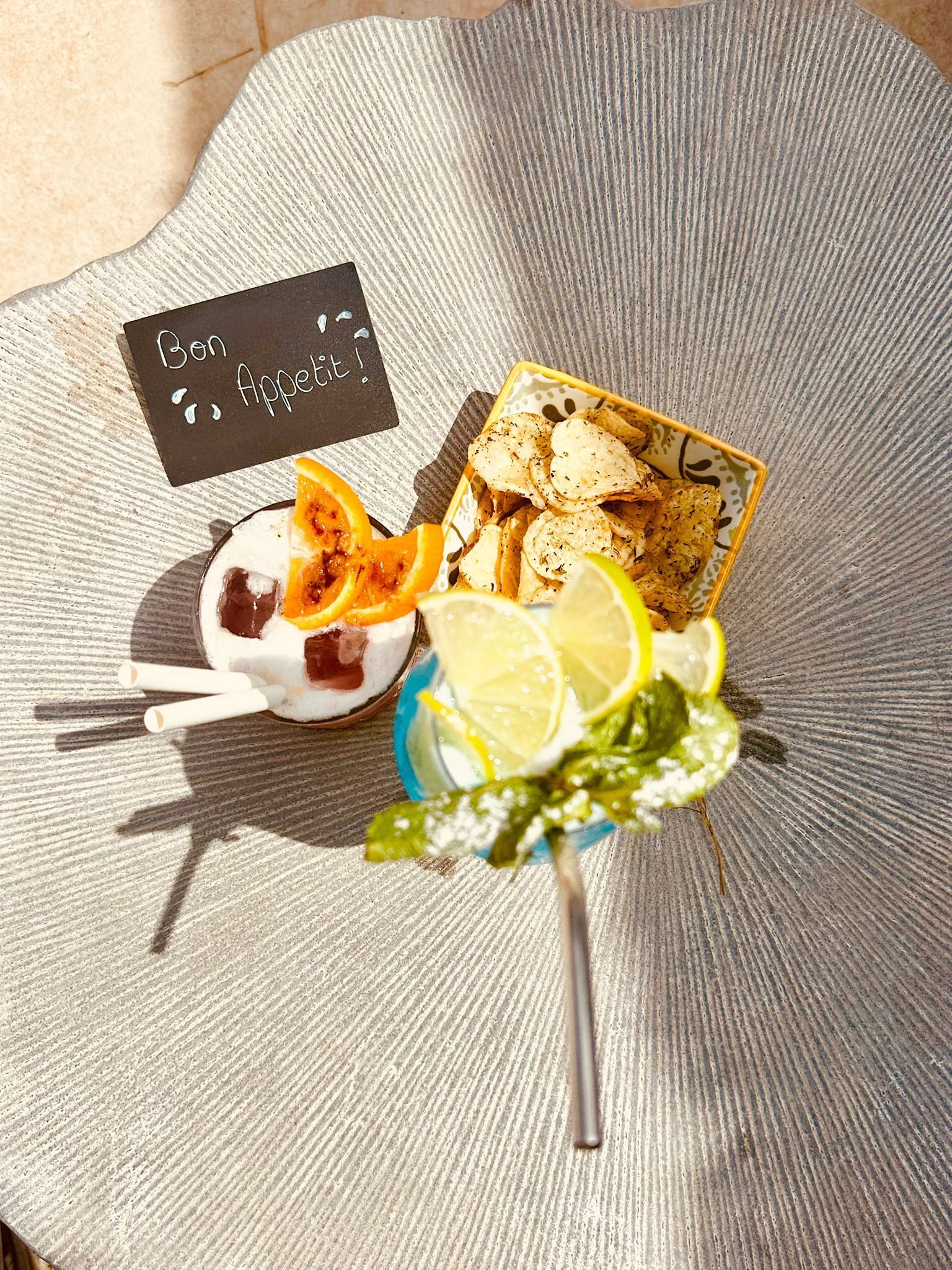 Preparación de alimentos con un tazón de yogur coronado con rodajas de naranja, un plato de papas fritas y una bebida con rodajas de limón y hojas de menta.