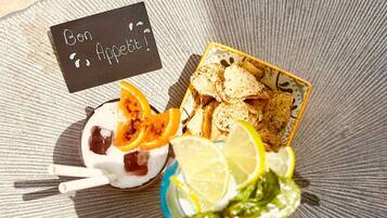Preparación de alimentos con un tazón de yogur coronado con rodajas de naranja, un plato de papas fritas y una bebida con rodajas de limón y hojas de menta.