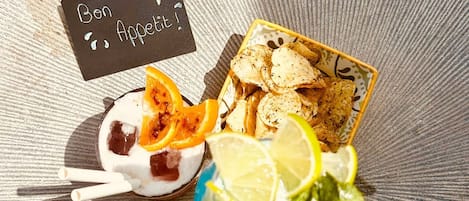 Un repas composé d’un bol de yaourt garni de tranches d’orange, d’une assiette de chips et d’une boisson contenant des tranches de citron et des feuilles de menthe.