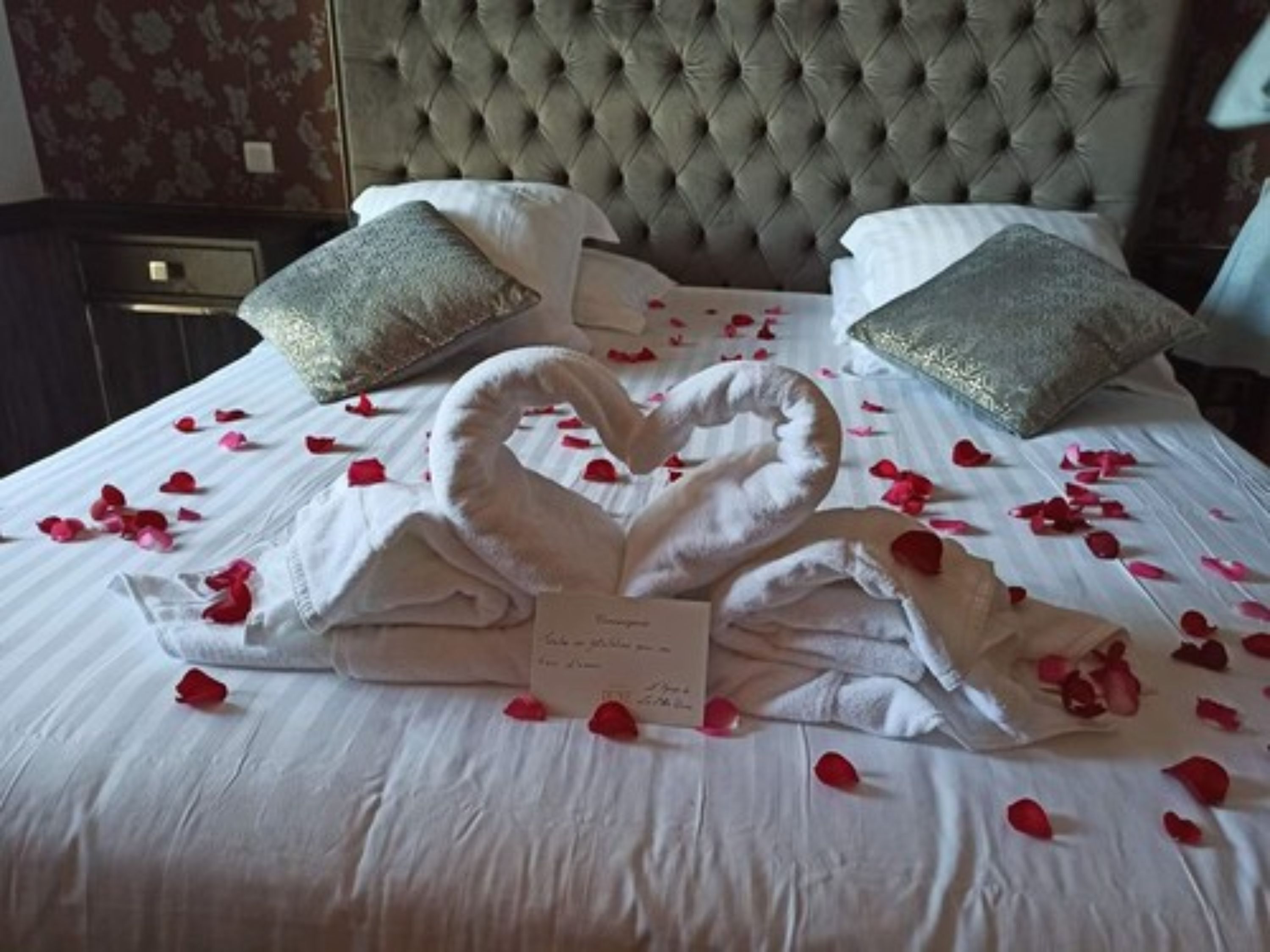 A bed with a heart-shaped towel arrangement and rose petals, a tufted headboard, and two decorative pillows.