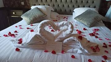 Una cama con una toalla dispuesta en forma de corazón y pétalos de rosa, un cabecero capitonado y dos almohadas decorativas.
