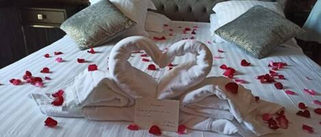A bed with a heart-shaped towel arrangement and rose petals, a tufted headboard, and two decorative pillows.