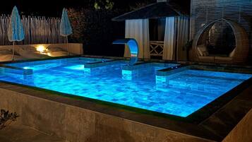 Una piscina con diseño geométrico, rodeada por un solárium de piedra. Se observan dos estructuras con forma de sombrilla y un pequeño pabellón con techo de paja.