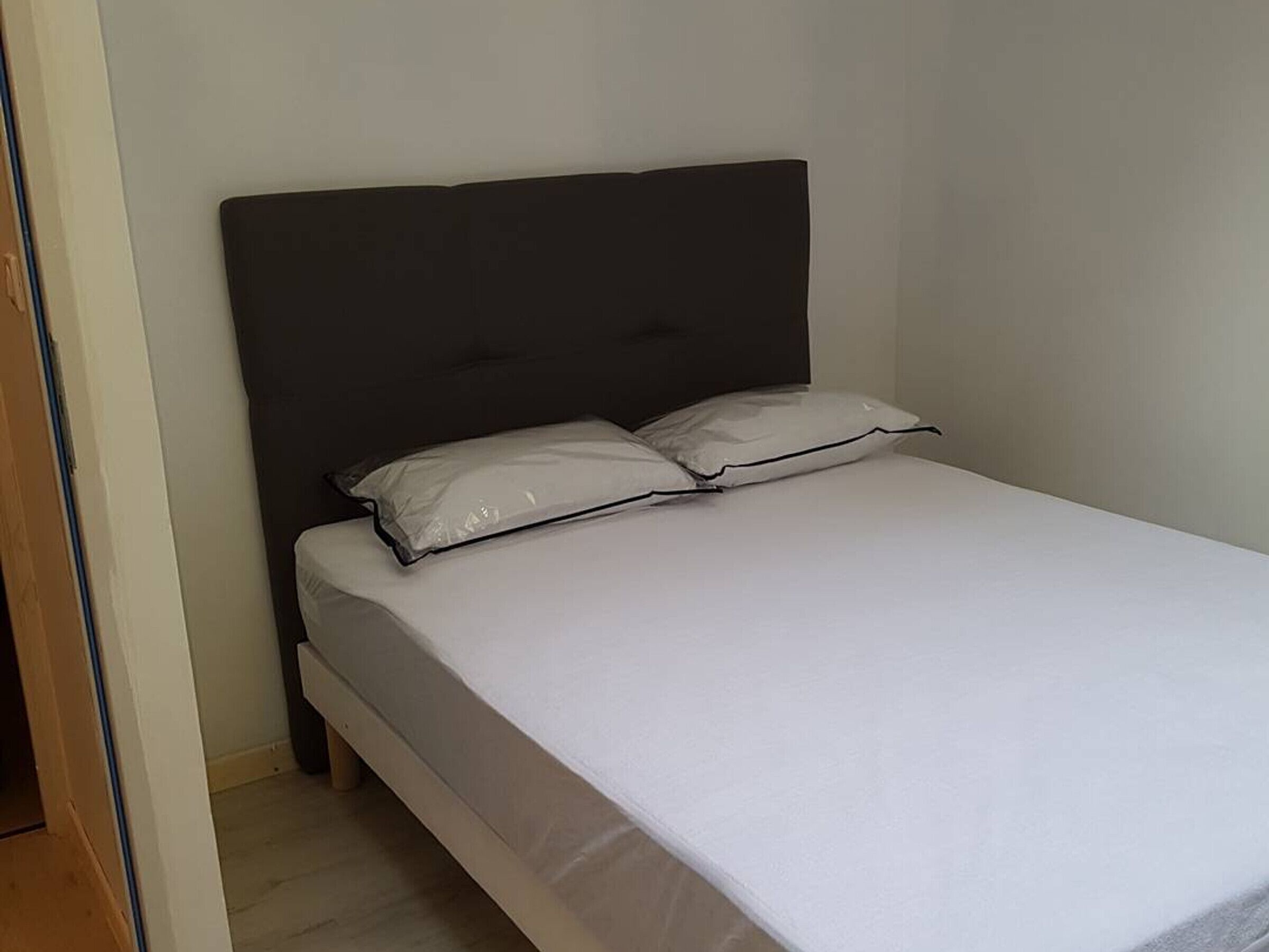 Un letto singolo con una testiera scura e due cuscini, in una stanza con pareti spoglie e pavimento in legno.