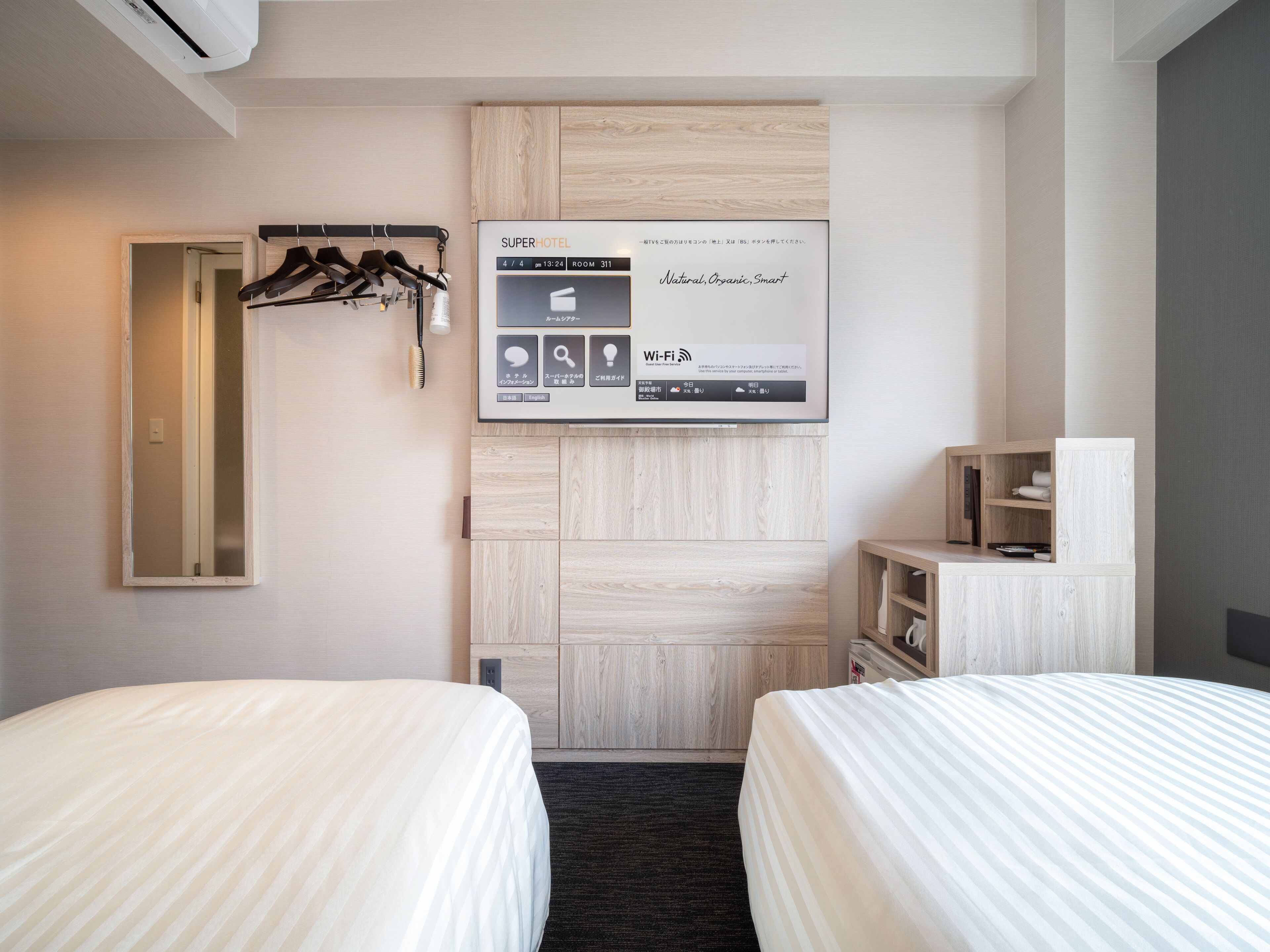 Quarto de hotel com duas camas, um guarda-roupa, um espelho e um painel digital.