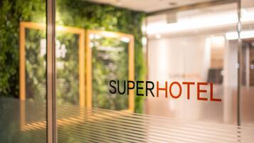 Eine Glastür mit dem Schriftzug „SUPERHOTEL“ und Blick auf eine grüne Wand sowie ein modernes Interieur.