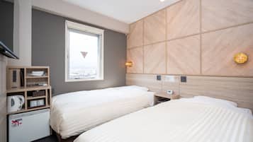 Quarto de hotel com duas camas, uma escrivaninha embutida, mini-frigorífico e janela com vista para a cidade.