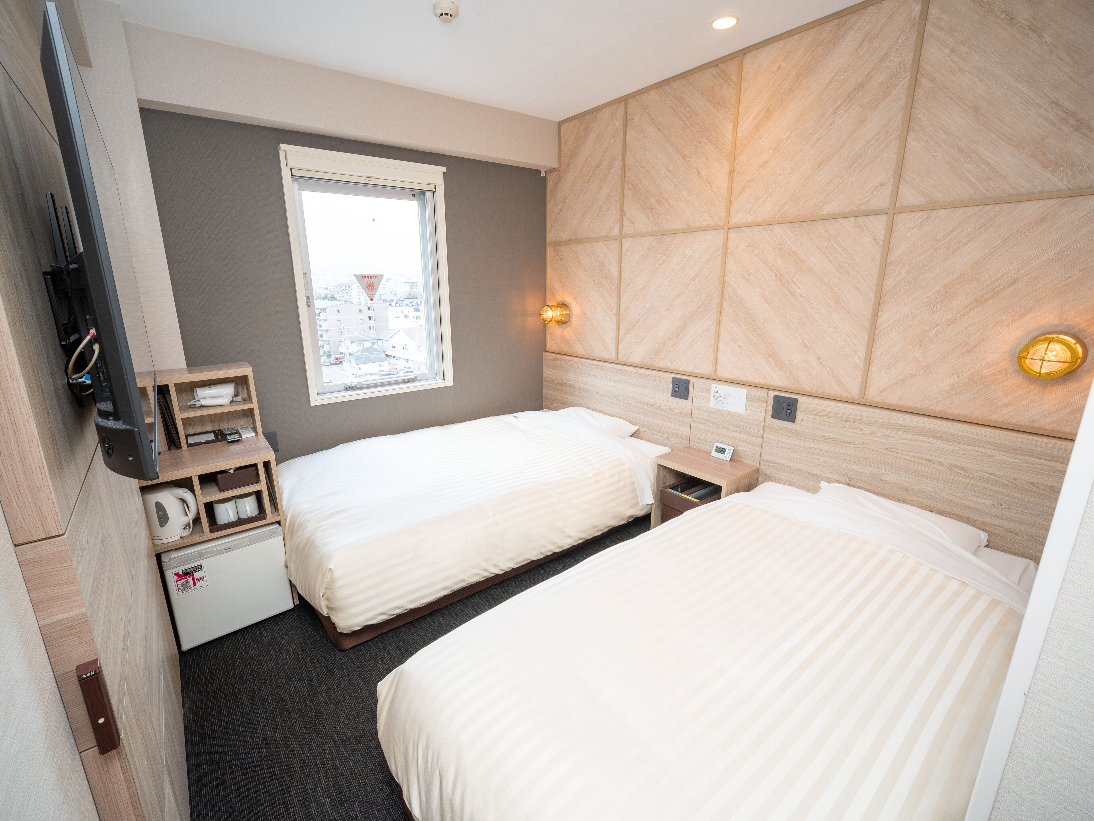 A hotel room with two beds, a window with a view of buildings, a wooden headboard, a bedside table with a clock, and a small shelf with a kettle and cups.
