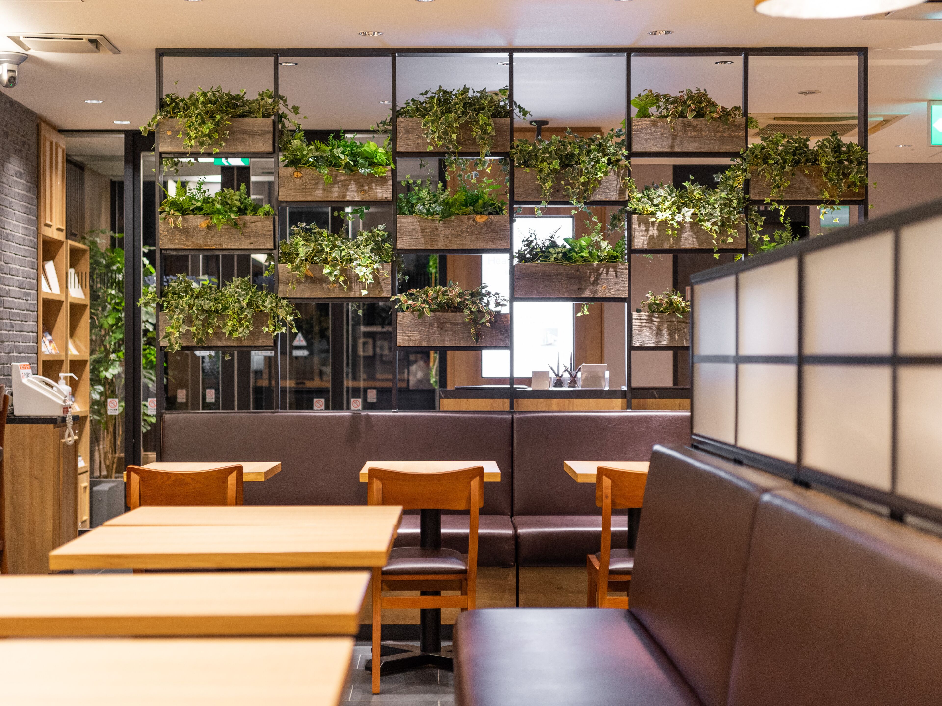 Et restaurantinteriør med træborde og bænke, en murstensvæg på den ene side, og en væg med træplantere i grønne planter.