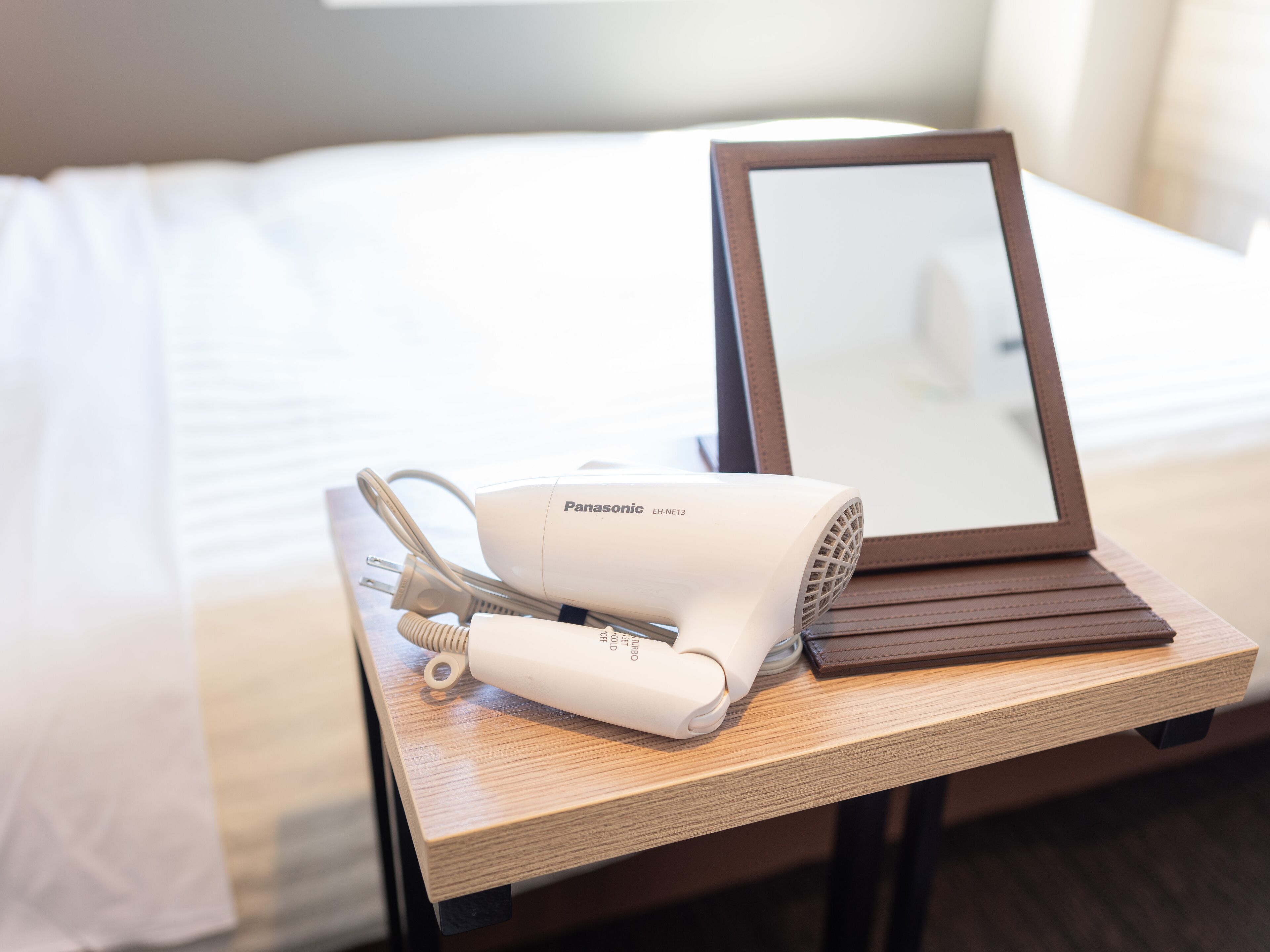 Un secador de pelo Panasonic EH-NE13 y un espejo sobre una mesita de noche en una habitación de hotel.