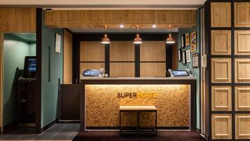 A reception desk with a wooden panel design and the name "SUPERHOTEL" prominently displayed. There are three pendant lights above the desk, and a small bench is placed in front of it.