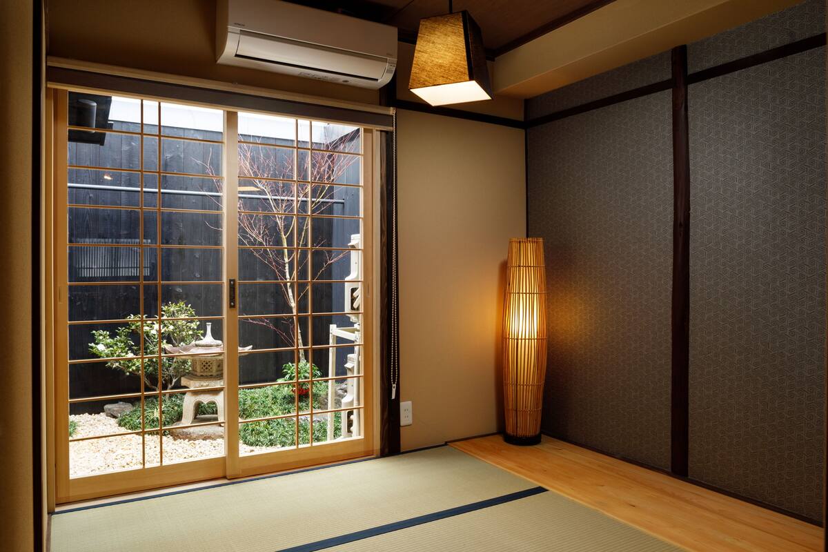 A traditional Japanese room with tatami flooring, a sliding door with a garden view, a bamboo lamp, and a wall-mounted air conditioner.