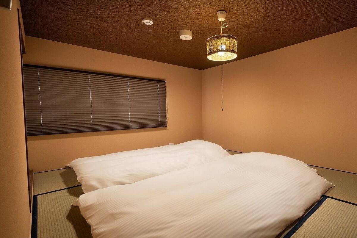 A room with two beds, a window with blinds, a ceiling light fixture, and tatami mats on the floor.